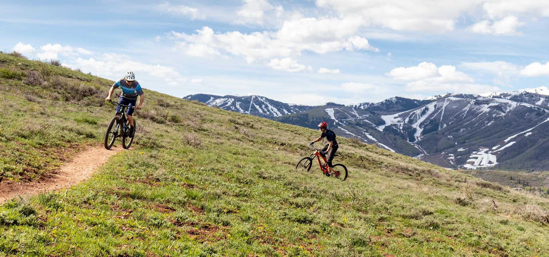 Jans Experts ride on a mountain bike trail with snow on the mountains in the background near Park City, UT.