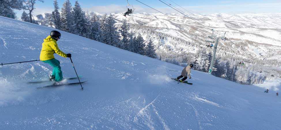 Two skiers ski down a run at Deer Valley Resort with a view of Park City, Utah.