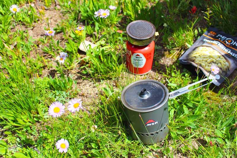  A backpacking stove setup sitting on the grass next to a freeze-dried backpacker meal.