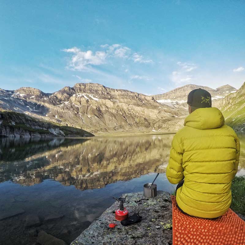 A camper eating a backcountry meal next to a lake in the mountains. Image courtesy of Nemo.