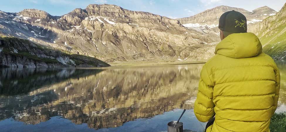 A camper sits on their sleeping pad next to a lake and prepares breakfast in the backcountry with mountains in the distance. Image courtesy of Nemo.