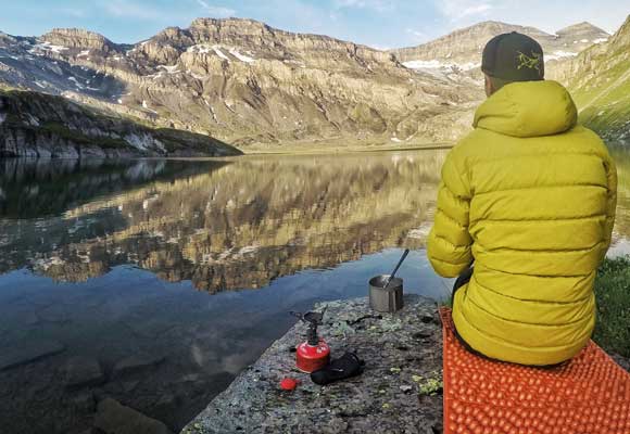 A camper sits on their sleeping pad next to a lake and prepares breakfast in the backcountry with mountains in the distance. Image courtesy of Nemo.