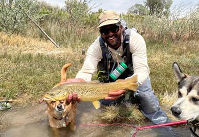 Jans fly fishing guide, Sam Elam, poses with two dogs while he holds a large brown trout caught in a river near Park City, UT